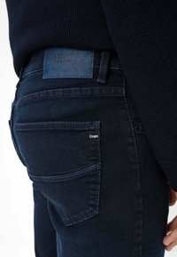 Jeans in denim blu scuro con una toppa in pelle del marchio, cuciture a contrasto e un classico design a cinque tasche. Texture liscia visibile.