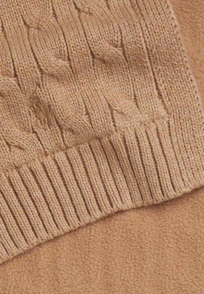 Beige kabelstickad tröja med ribbad nederkant och mjuk fleece-känsla i matchande beige. Har ett subtilt emblem på stickningen.