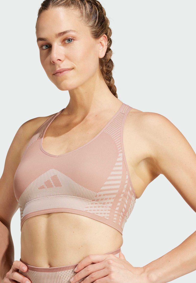 Sostén deportivo de color rosa claro, hecho de tela texturizada. Presenta un patrón geométrico, un diseño de espalda de nadadora y un logo en la banda.
