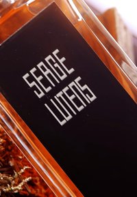 Flacone di profumo in vetro con un'etichetta nera e testo bianco. Il liquido all'interno è di colore ambra e il flacone ha una forma rettangolare.