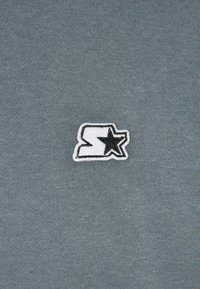 Grauer Baumwollstoff mit weicher Textur, der ein weißes und schwarzes gesticktes Logo enthält, bestehend aus einem 'S' und einem Sternmuster.