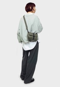Grå oversized sweatshirt lagdelt over en hvid skjorte, parret med løse mørke bukser. En lille olivengrøn crossbody-taske med en sort strop.