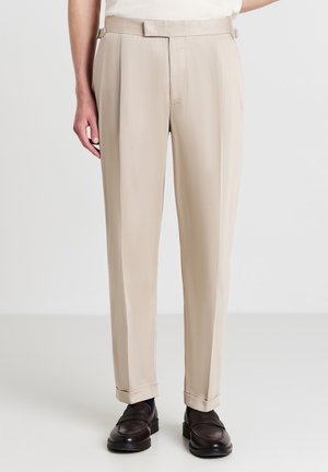 Beige maßgeschneiderte Hose mit geradem Schnitt, aus glattem Stoff gefertigt. Verfügt über seitliche Verstellmöglichkeiten und Bündchen an den Knöcheln, ergänzt durch dunkle Schuhe.