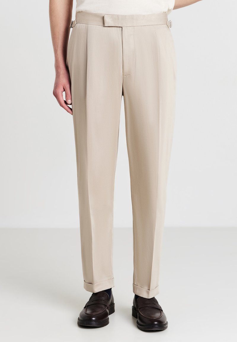 Beige maßgeschneiderte Hose mit geradem Schnitt, aus glattem Stoff gefertigt. Verfügt über seitliche Verstellmöglichkeiten und Bündchen an den Knöcheln, ergänzt durch dunkle Schuhe.
