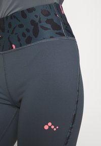 Vysoké sportovní legíny tmavě šedé barvy s páskem v černé a tyrkysové barvě, zdobeným růžovými akcenty, vyrobené z elastického materiálu.