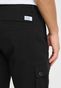 Pantalons cargo noirs en tissu texturé, avec une poche plaquée et des accents de boutons. Comprend une étiquette sur la poche arrière.