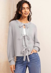 Cardigan tricotat gri, cu guler rotund, închidere cu cravată pe față, mâneci lungi și un design cropped. Asortat cu blugi albaștri.