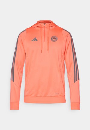 Narančasta dukserica s kapuljačom sivoj Adidas trakama i logom. Izrađena od glatkog materijala, sadrži patentni zatvarač i rebraste rukave i donji rub.