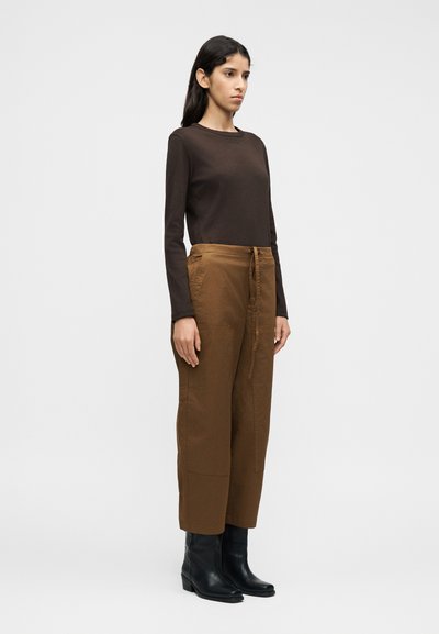 Καφέ πουκάμ ισο με μακριά μανίκια συνδυασμένο με καφέ παντελόνι cargo. Το παντελόνι έχει πλαϊνές τσέπες και μπροστινό δέσιμο. Το μοντέλο φοράει μαύρες μποτίνες αστραγάλου.