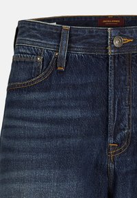 Mørkeblå denimjeans med oransje søm, standard fem-lomme design, metallbeslag og knappefeste. Merketagg synlig på livlinjen.