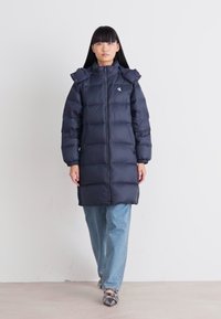 Kvinna klädd i lång svart pufferjacka med huva, ljusblå jeans och svarta spetsiga skor, ståendes mot en enfärgad vit vägg.
