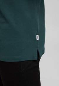 Ourlet de polo en tissu texturé bleu sarcelle avec petite fente latérale et étiquette blanche de la marque Esprit, porté sur un pantalon noir.