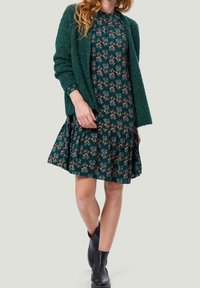 Vestido floral verde con dobladillo rizado, llevado bajo un cárdigan verde texturizado; combinado con botas negras hasta el tobillo. Presenta mangas largas y un cuello redondo.