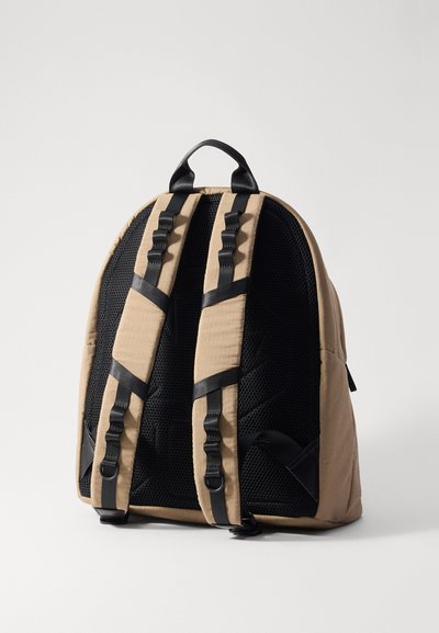 Khaki-Rucksack mit schwarzem Netzrücken, ausgestattet mit verstellbaren Riemen, schlankem Design und einem Griff. Strukturierte Akzente entlang der Riemen.