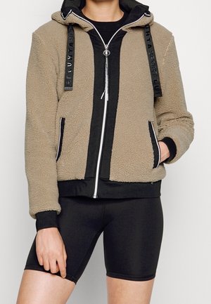Persoon draagt een beige fleece jas met ritssluiting, zwarte bies en capuchon, over zwarte fietsbroek, met één hand in de jaszak, tegen een effen achtergrond.