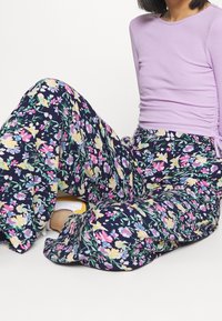 Person som sitter i korslagda ben, iklädd en lavendelfärgad långärmad topp och marinblå blommiga vida byxor med vita sneakers.