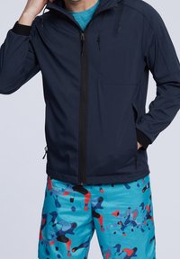 Námořnická modrá zipová bunda s vysokým límcem, dvěma bočními kapsami a nastavitelnými šňůrkami; v kombinaci s barevnými vzorovanými plavkami.