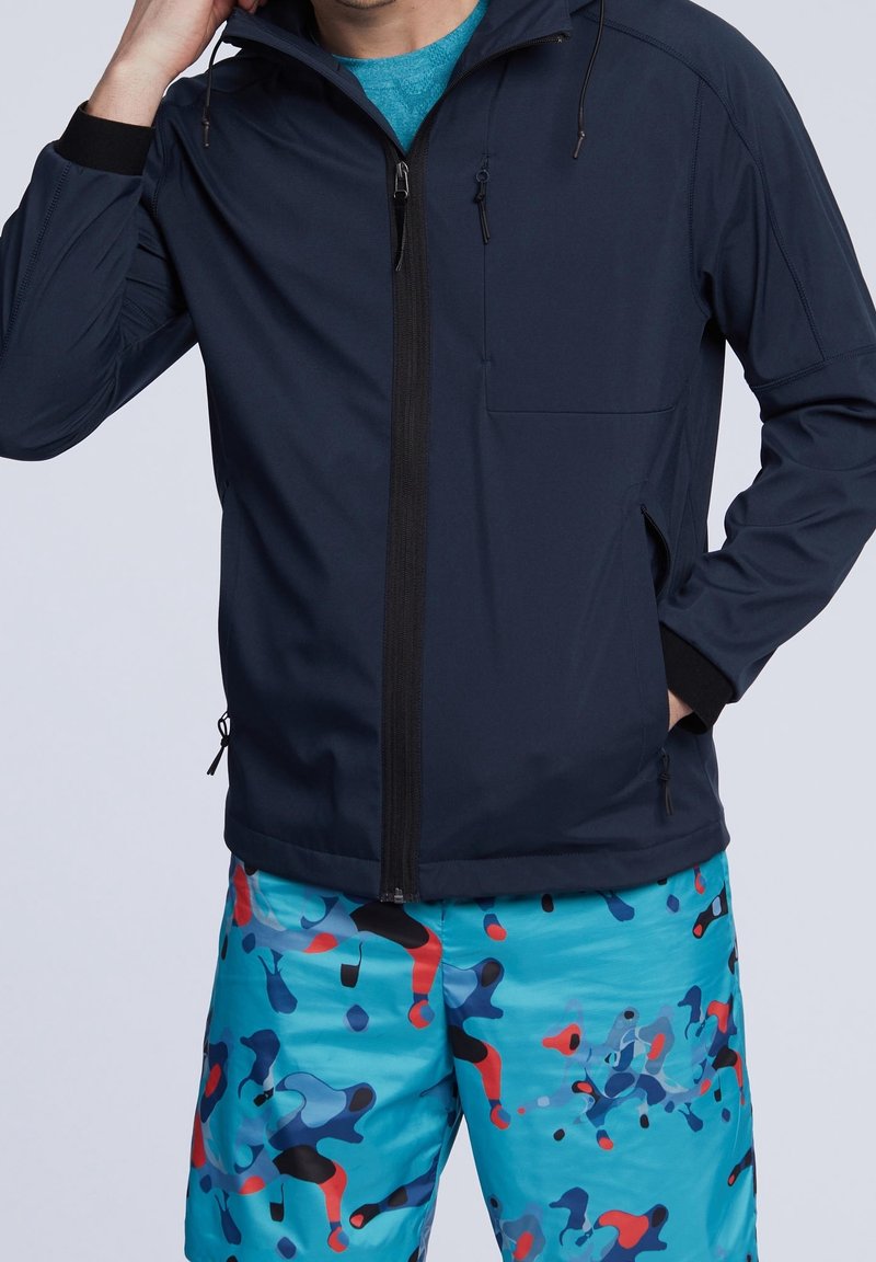 Námořnická modrá zipová bunda s vysokým límcem, dvěma bočními kapsami a nastavitelnými šňůrkami; v kombinaci s barevnými vzorovanými plavkami.