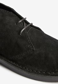 Black suede shoe with a smooth texture, featuring gray laces, detailed stitching, and a round toe design. Rubber sole visible at the bottom.