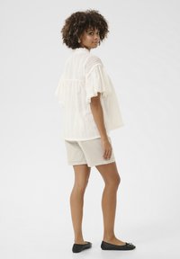Blusa bianca e strutturata con maniche a volant, abbinata a pantaloni corti beige chiaro e ballerine nere. Nessun motivo o hardware visibile.