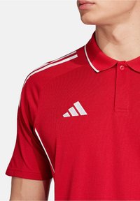 Czerwona koszulka polo wykonana z teksturowanej tkaniny, z białym wzorem trzech pasków na ramionach i białym logo Adidasa na klatce piersiowej.