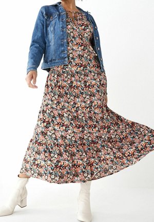 Woman wearing a floral maxi dress with a blue denim jacket and white heeled boots, dress flowing outward as she moves.