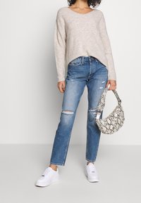 Kvinna som bär en beigefärgad stickad tröja och rivna blå jeans, håller en handväska med ormskinnsmönster och har vita sneakers.