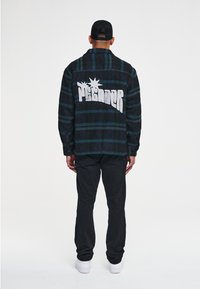 Czarna i zielona flanelowa koszula w kratę z dużym białym nadrukiem na plecach. Do tego czarne spodnie i białe trampki.