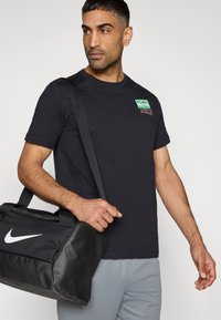 Μαύρο T-shirt με κοντά μανίκια και πράσινο κόκκινο λογότυπο στο στήθος, συνδυασμένο με γκρι παντελόνι. Κρατείται μια μαύρη τσάντα γυμναστηρίου με λευκό λογότυπο Nike.