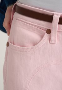 Jeans in denim rosa con superficie testurizzata, caratterizzati da una tasca frontale a forma arrotondata, chiusa da un bottone marrone e una cintura marrone.