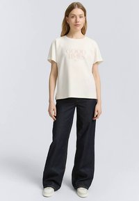 T-shirt crema a maniche corte con grafica "GOOD TIMES" in rosa tenue, abbinata a jeans larghi scuri e sneakers bianche.