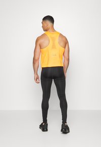 Camiseta de entrenamiento sin mangas para hombre en material de malla naranja brillante, con un diseño de espalda nadadora y panel trasero reflectante, combinada con leggins negros.