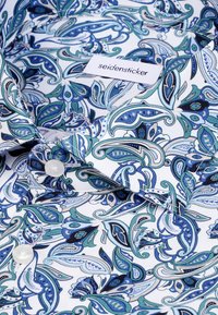 Geprinte blouse met een blauwgroen paisley-ontwerp op een witte achtergrond, met een puntige kraag en een witte knoopsluiting.