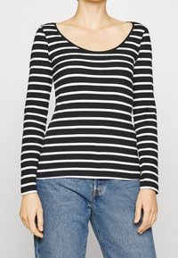 Frau trägt langärmliges Oberteil mit schwarzen und weißen horizontalen Streifen und blaue Jeans, Hände entspannt an den Seiten, vor schlichtem Hintergrund.