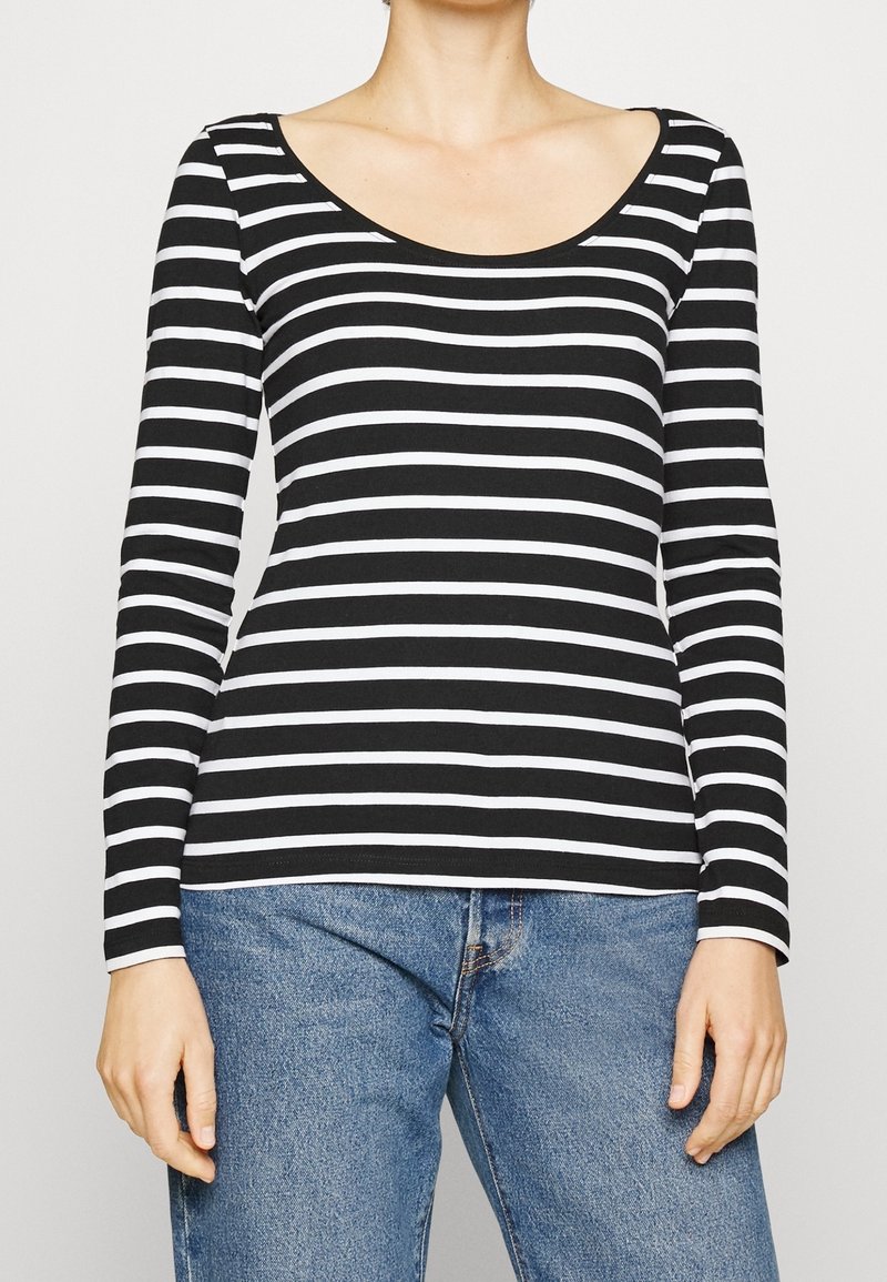 Frau trägt langärmliges Oberteil mit schwarzen und weißen horizontalen Streifen und blaue Jeans, Hände entspannt an den Seiten, vor schlichtem Hintergrund.