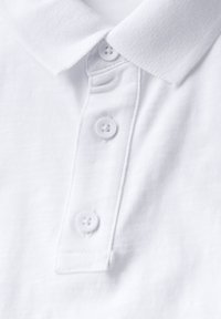 Bílá polo tričko s texturovanou tkaninou, klasickým límcem a třemi bílými knoflíky podél lišty pro zapínání.