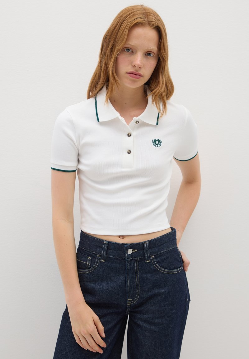 Polo shirt bianco a maniche corte, con trim verde e logo. Placket con bottoni. Abbinato a jeans blu scuro con cuciture dorate.