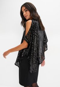 Capa nera con paillettes e spalle scoperte, caratterizzata da un design fluido e una texture morbida. Indossata sopra un semplice vestito nero.