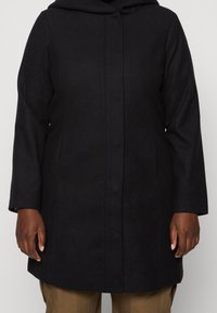 Schwarzer Langarmmantel mit verdeckten Knöpfen, getragen von einer Person mit braunen Hosen, die Hände entspannt an den Seiten, vor einem neutralen Hintergrund.