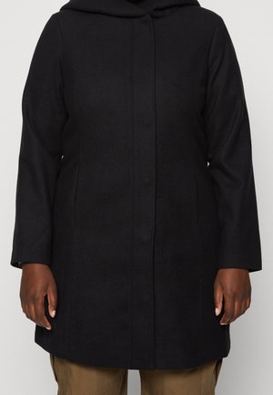 Svart långärmad kappa med dolda knappar som bärs av en person med bruna byxor, händerna avslappnat vid sidorna, mot en neutral bakgrund.