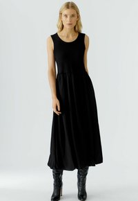 Schwarzes ärmelloses Kleid, lockere Passform mit einem ausgestellten Rock und rundem Ausschnitt, aus glattem Stoff; kombiniert mit schwarzen Stiefeln bis zum Knie.