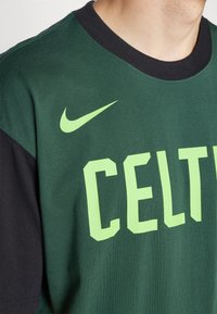Camiseta de algodón verde con mangas cortas negras. Presenta un texto en negrita color verde claro "CELTIC" y un logo de Nike en verde claro en el frente.
