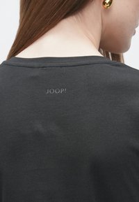 Crna majica glatke teksture, s okruglim izrezom i logotipom "JOOP!" u sivom tisku na stražnjoj strani. Minimalistički dizajn.