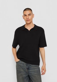 Must have lühikeste varrukatega must polo-särk, millel on sooniktekstuur ja nurgeline krae, koos helesiniste denim-pükstega.