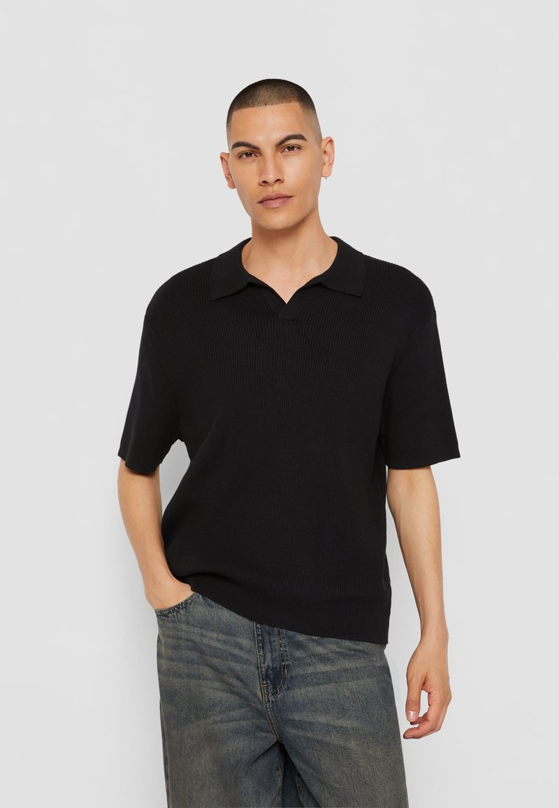 Must have lühikeste varrukatega must polo-särk, millel on sooniktekstuur ja nurgeline krae, koos helesiniste denim-pükstega.