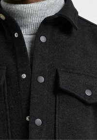 Svart ulljacka med texturerad yta, med tryckknappsstängning, och en grå ribbad polokrage under. Fickor syns på jackan.