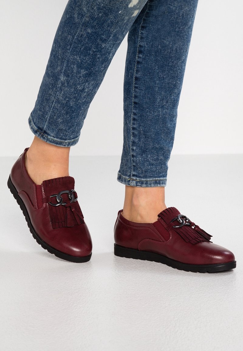 Chaussures en cuir bordeaux avec une semelle plate, présentant un accent à franges et à chaîne sur le devant. Portées avec un jean bleu retroussé.
