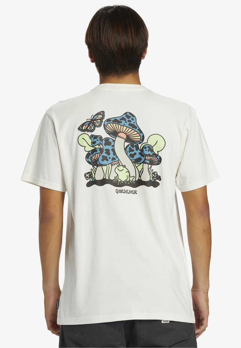 Personne portant un t-shirt blanc avec un motif coloré de champignons bleus, une grenouille verte et un papillon dans le dos, se tenant de dos.