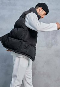 Gilet rembourré noir avec un col montant, design matelassé, fermeture éclair latérale et coupe ample, porté sur un haut texturé blanc et un pantalon.