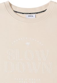 T-shirt beige à col rond avec un texte composé de petits points disant "SUNKISSED SOUL", un petit palmier et les phrases "EMBRACE CHANGE" et "TAKE A BREAK FEEL JOY."